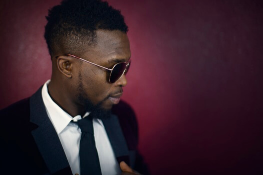 man with beard wearing black suit and sunglasses with a purple backdrop