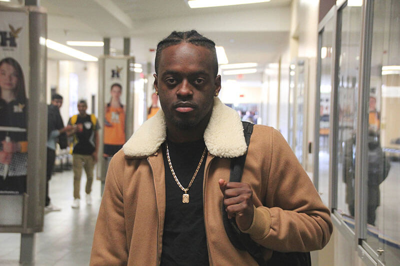 Student in jacket standing in hall with hand on backpack strap.