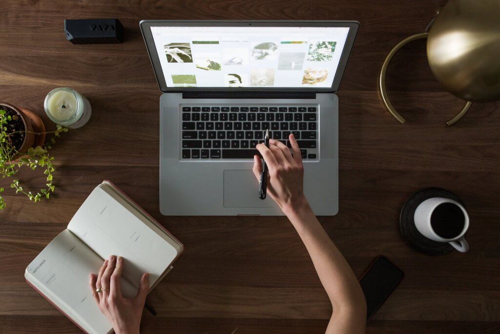 Hands reaching towards a laptop and notebook on a wooden table, with coffee and plant on the table
