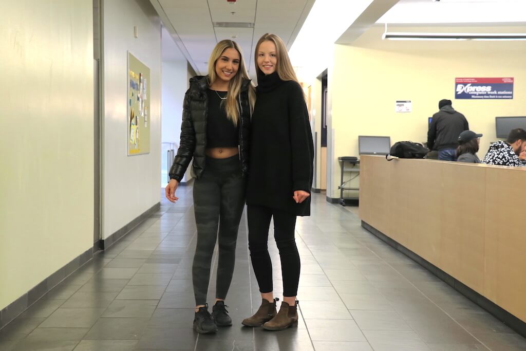 Girl wearing cropped jacket, with t-shirt, tights and running shoes standing next to another girl wearing oversized turtle neck sweater, tights and boots.