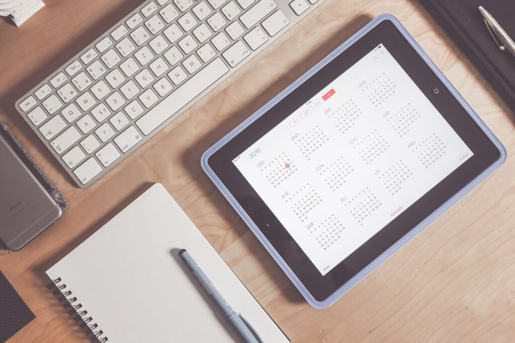 A tablet with a calander open on a wooden desk next to a keyboard and notebook