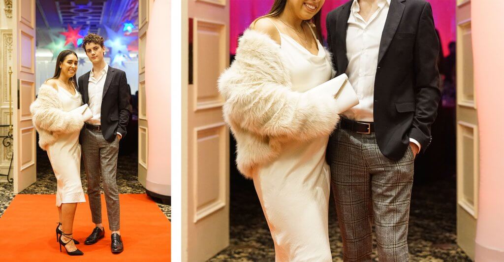 A young woman wearing a dress with a fur jacket, and young man wearing a suit pose at Gala