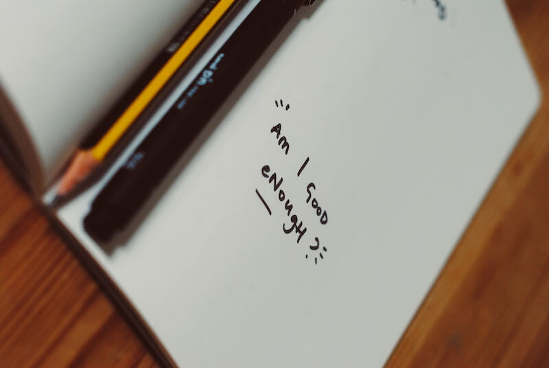 Notebook that reads "am I good enough?" with pen and pencil beside it