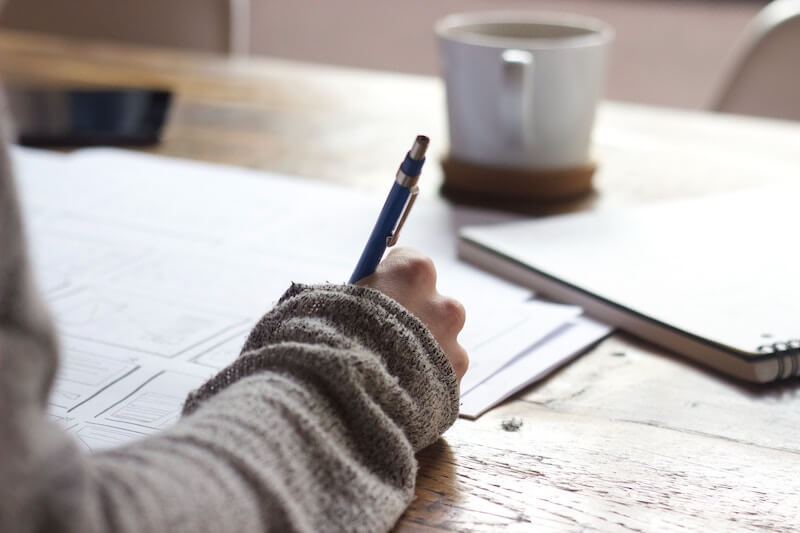 close up of arm writing on notebook with a mug in the background