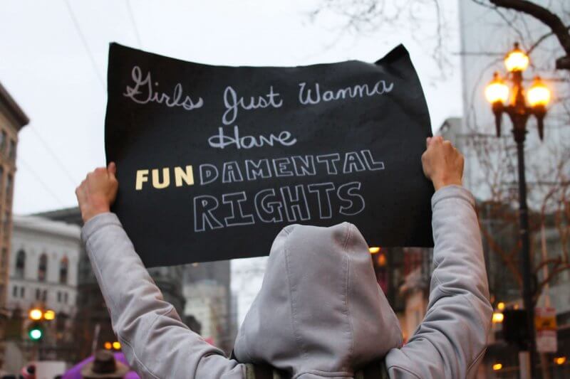 Person in hoodie holding black sign that reads "Girls just wanna have FUNdamental rights"