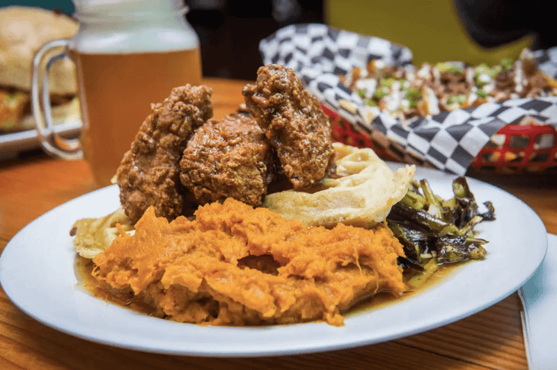 Vegan chicken and waffles, collard greens and sweet potatoes on plate with basket of nachos and mug of beer in background.