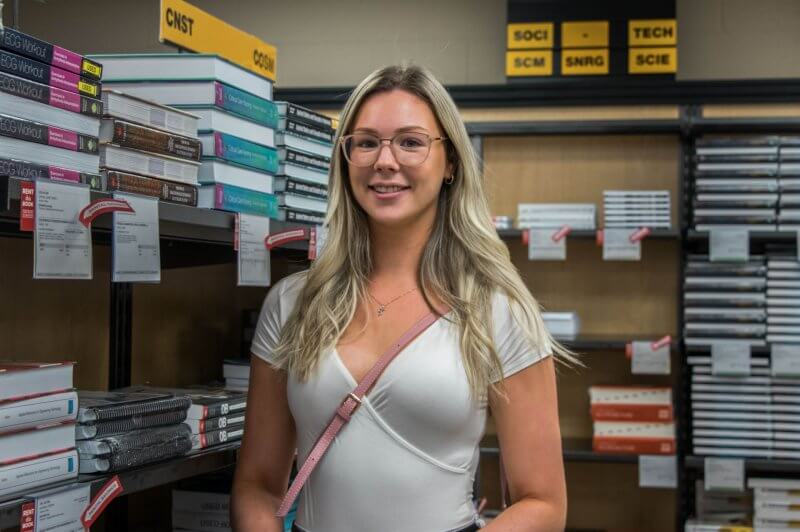 blonde girl wearing white shirt and glasses in front of bookshelf