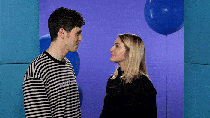 A man and a woman stand next to each other. The woman blows glitter into the air and disappears; the man looks into the camera, confused.