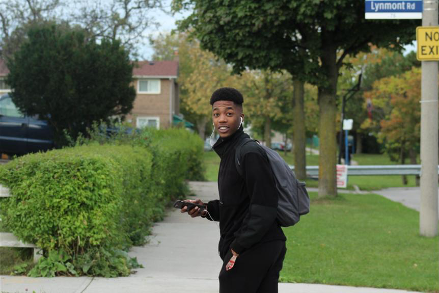 IGNITE 2020-2021 Board of Directors member Ryan Stafford wears a black jacket and a grey backpack while standing outside on a sidewalk. To his left, there are a row of bushes. To his right, there are oak trees. Stafford holds a smartphone in his right hand and looks over his left shoulder at the camera.