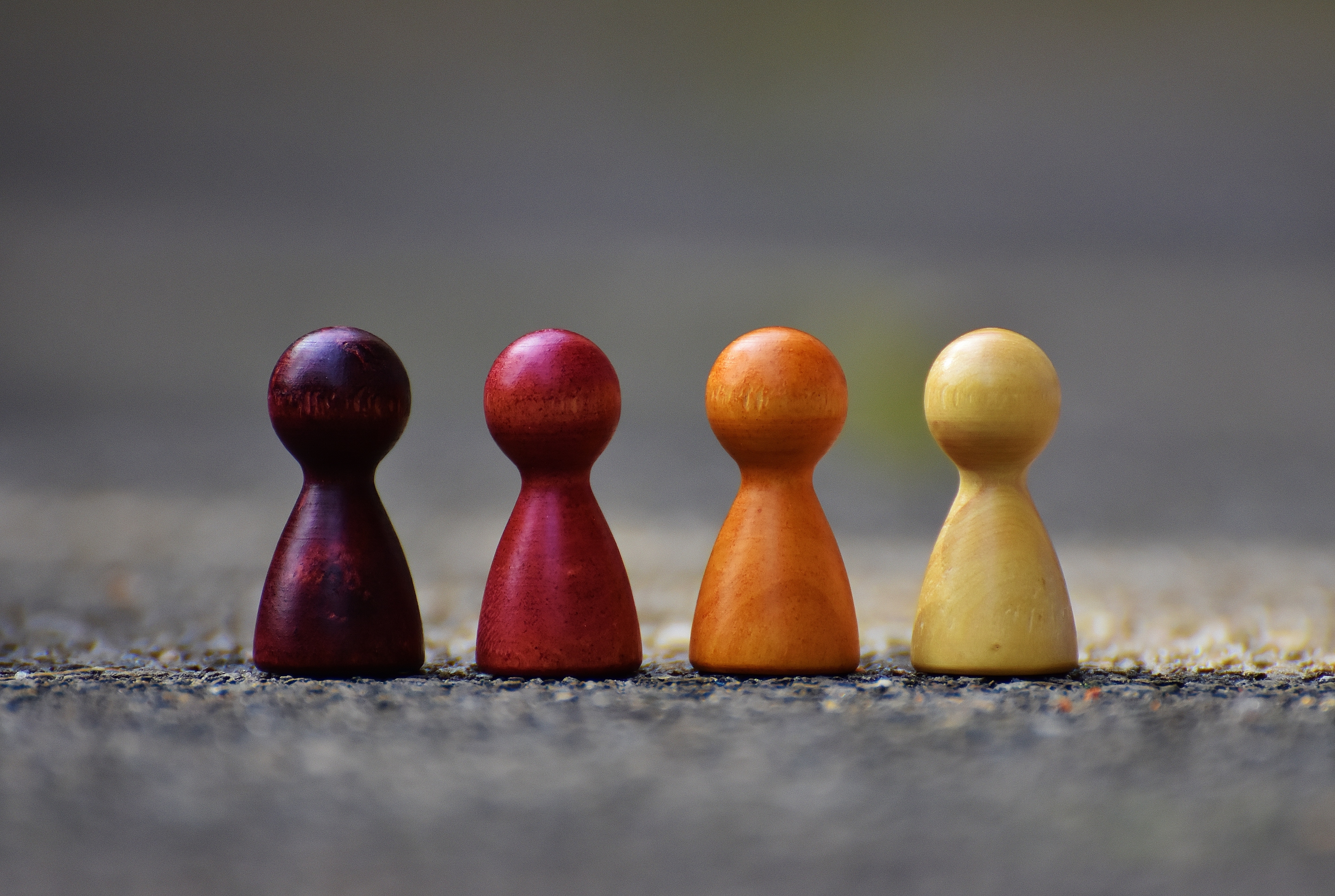 Four humanoid wooden board game pieces sit in a line on a cement ground. From left to right, they are purple, red, orange and yellow.