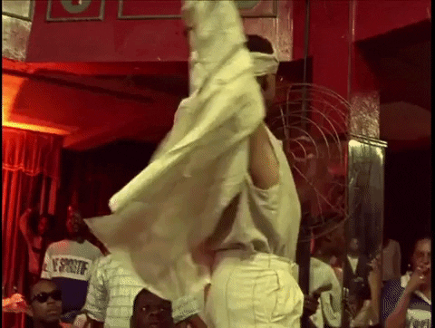 A young black person in a monochrome cream-coloured outfit holds his arms above his head and twists his torso, dancing.