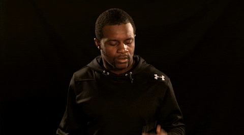 A man in a black Under Armour sweater holds up a cell phone and appears shocked.