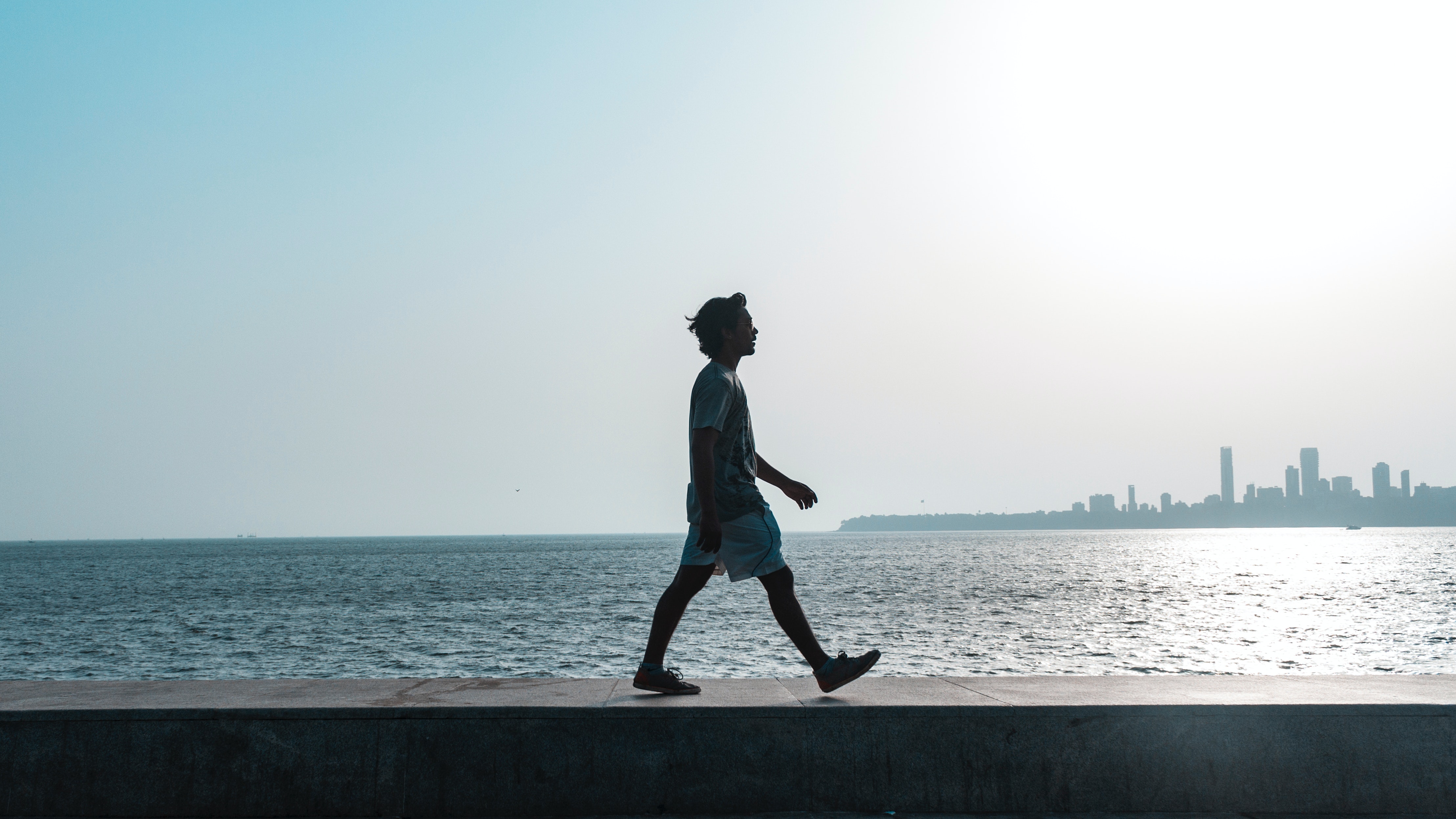 silhouette of a young person walking alone on a pier. Behind them is a still body of water and a cloudless blue sky.