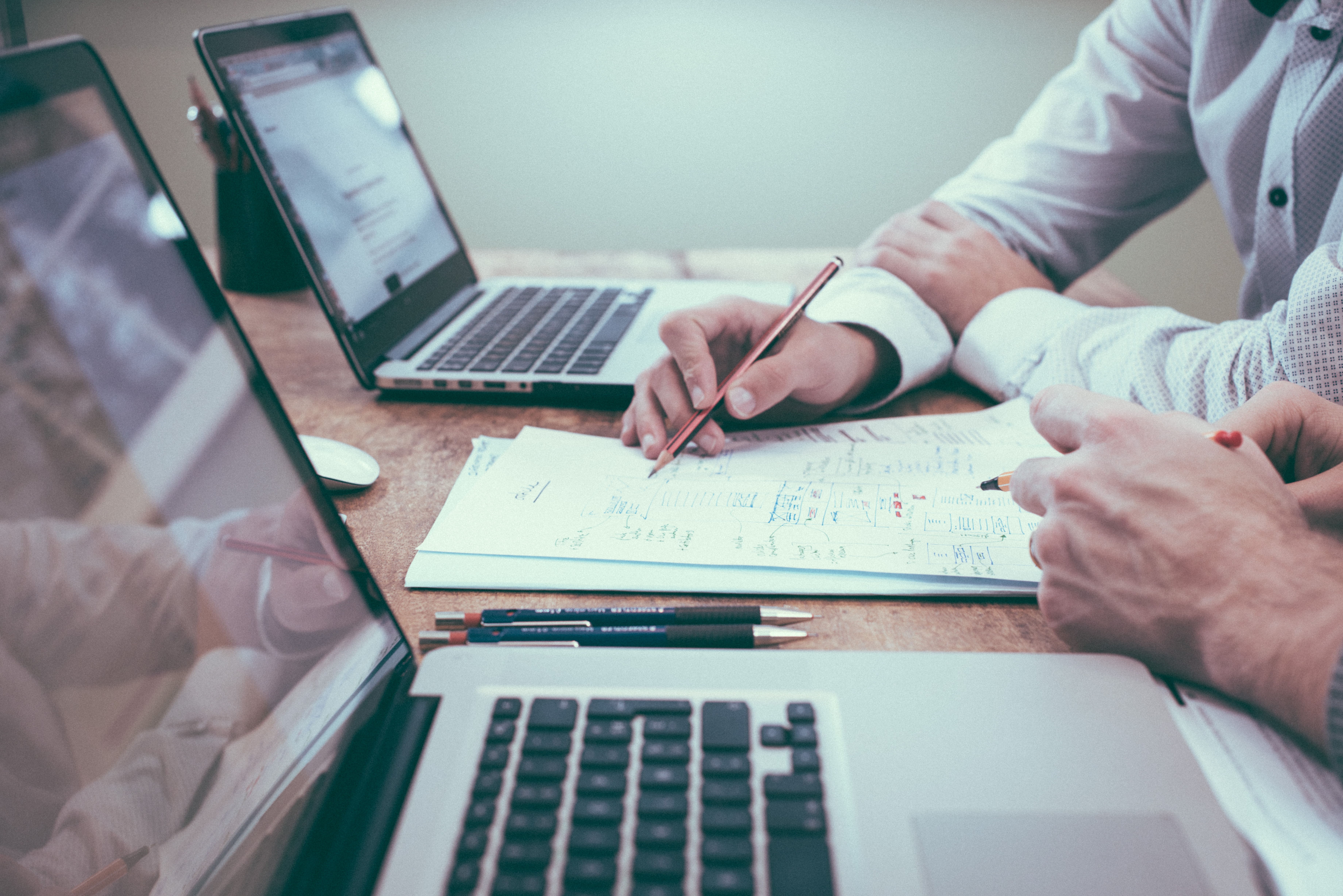 side view of two people's hands as they sit at a desk in front of two MacBook laptops and paperwork.