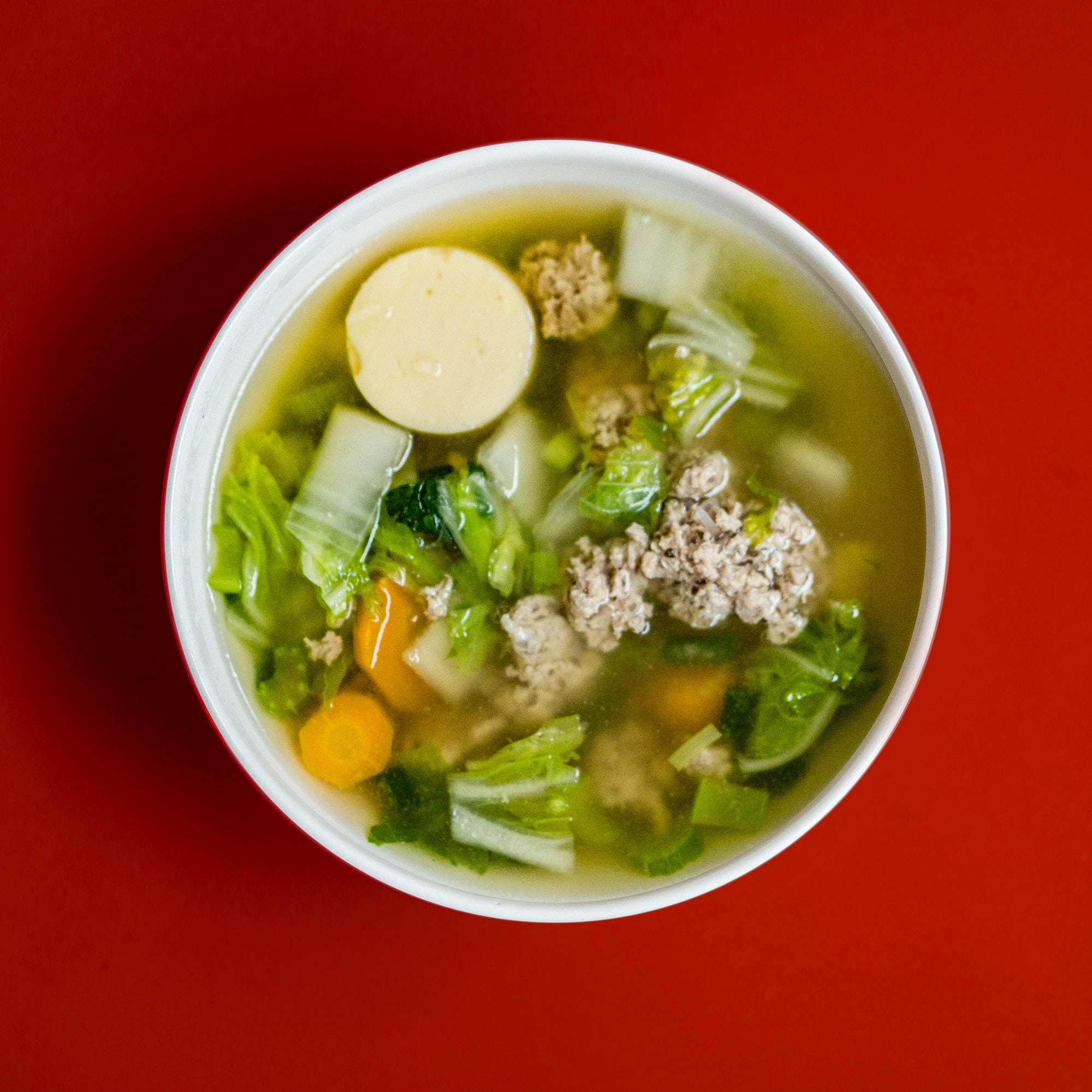 A bowl of meat and vegetable soup in a white bowl on a red background.