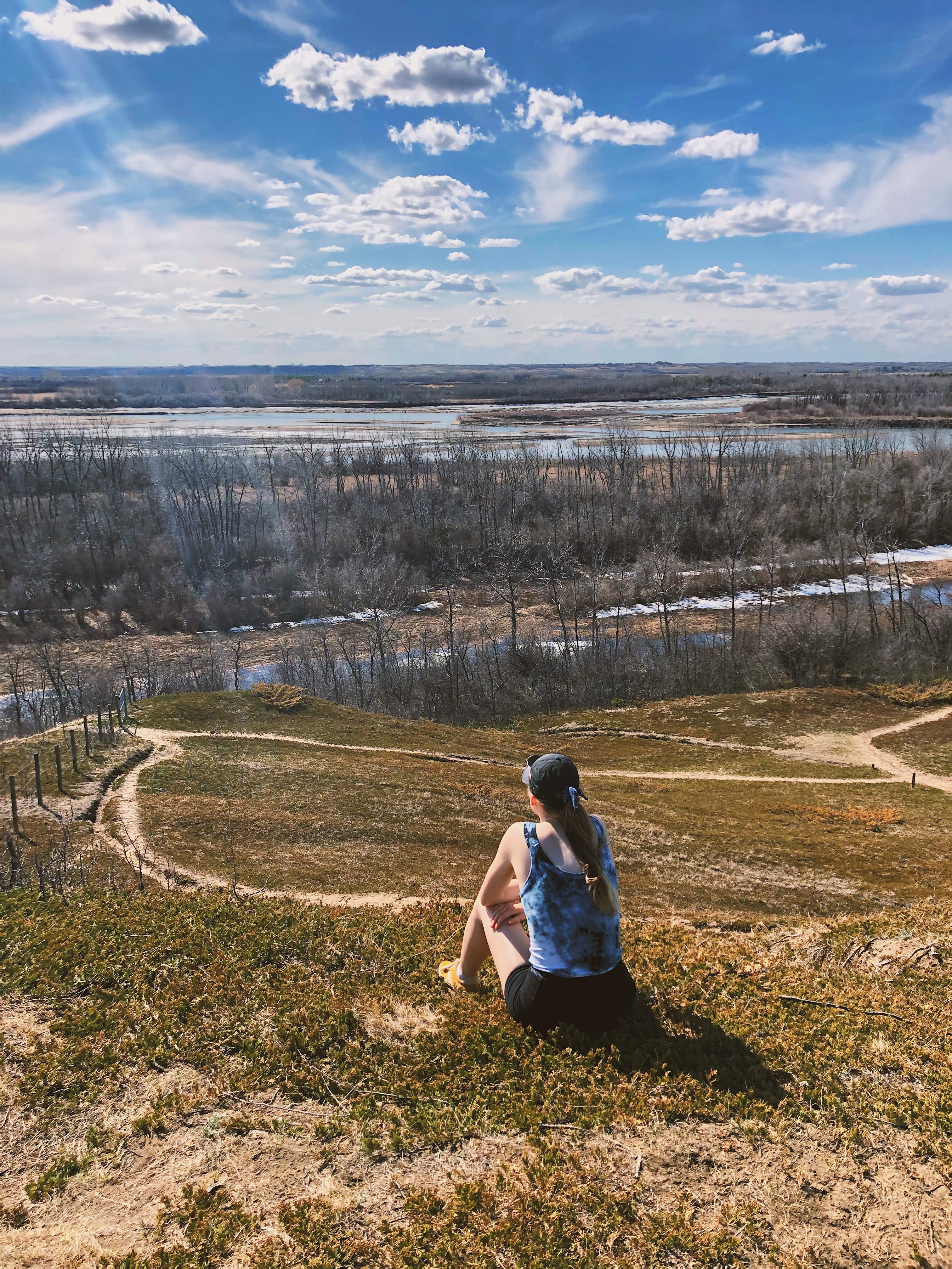 IGNITE content writer Gabby Dumonceaux sits facing away form the camera overlooking a sunny prairie landscape. She wears black shorts, a black ball cap, and a blue-and-white tank top. Her straight blonde hair is in a ponytail. In front of her are hills of brown and green grass accentuated with foot paths made of sand. Leafless trees stand in a row along the horizon in front of a shallow lake.