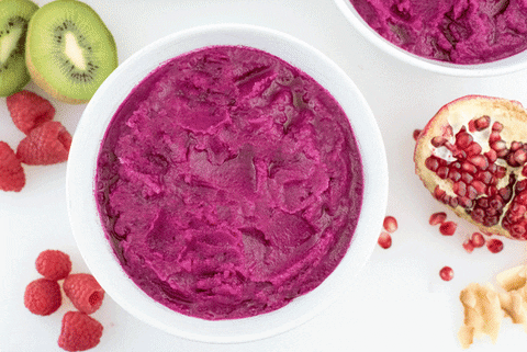 Stop-motion animation of garnishes being added to a deep pink smoothie bowl. The garnishes include sliced bananas, sliced kiwis, mango puree, raspberries, pomegranate seeds and chia seeds.