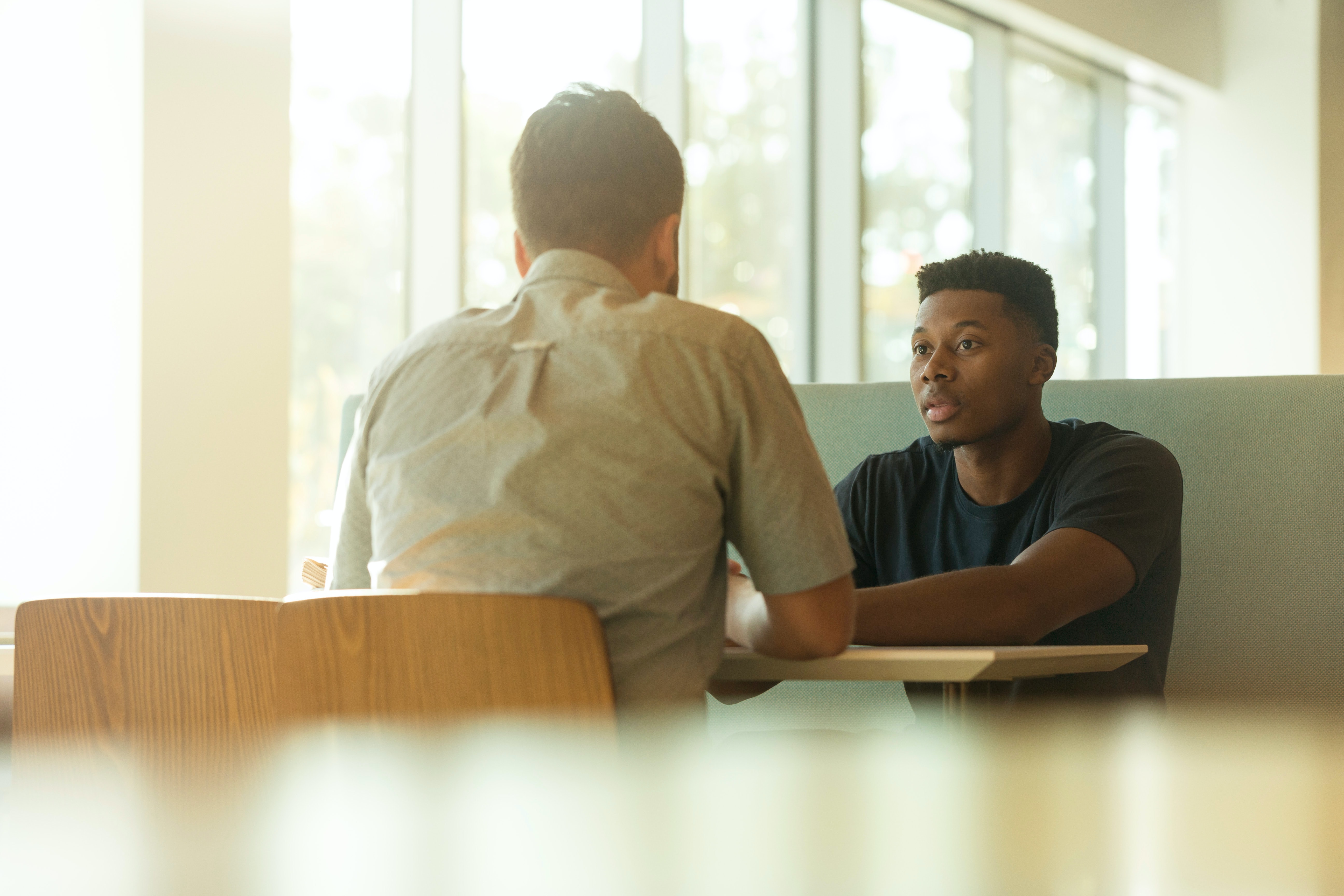 Two men sit across a table, talking.