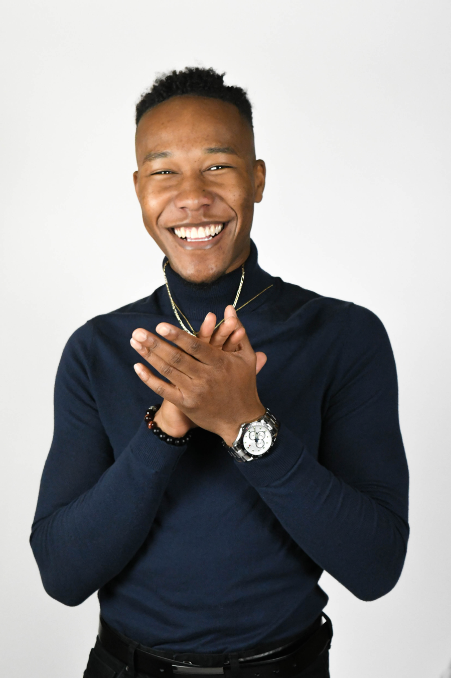 IGNITE 2020-2021 Board of Directors member from the University of Guelph-Humber, Stefan Thomas, wears a black turtleneck sweater, a gold chain on his neck, and a watch on his left wrist. His hands are clasped below his chin and he smiles at the camera.