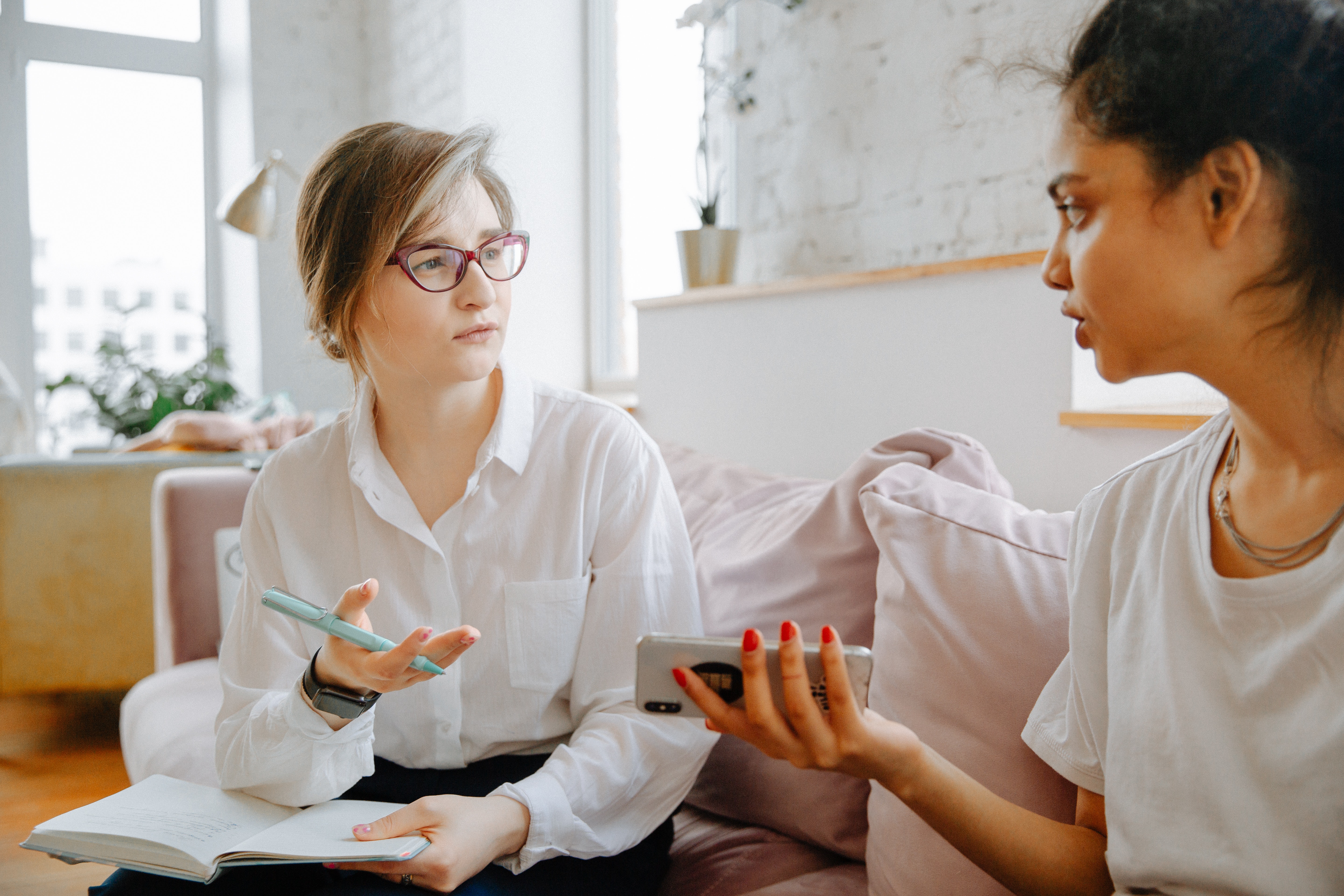 Two women converse on a pale pink couch. On the left, a woman with light blonde hair in a ponytail and red glasses wearing a white blouse gestures with her right hand. On the right, a dark-haired woman wearing red nail polish holds an iPhone in her left hand.