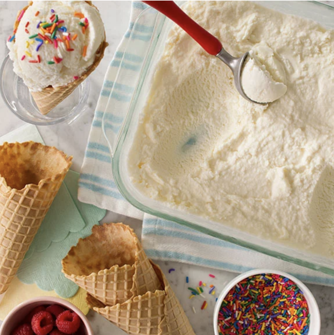 A glass dish of homemade vanilla ice cream surrounded by an array of waffle cones and colourful sprinkles.