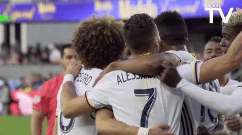 A soccer team huddles together with their arms around one another. A player joins the huddle by placing his arms around his teammates.