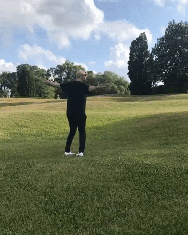 A bald man wearing black pants, a black shirt, and white shoes spins around with his arms extended in an empty green field.