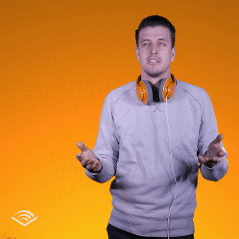 A man wearing a grey sweater with orange headphones around his neck shrugs his shoulders indecisively.